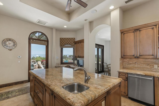 kitchen featuring a center island with sink, sink, stainless steel dishwasher, light stone counters, and ceiling fan