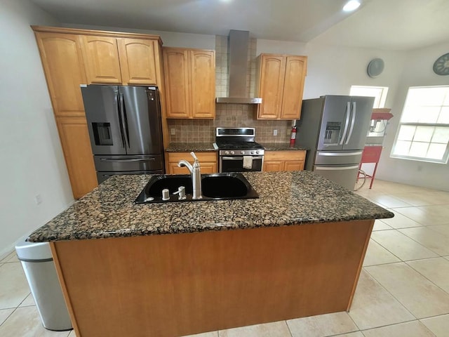 kitchen featuring appliances with stainless steel finishes, an island with sink, sink, dark stone countertops, and wall chimney range hood