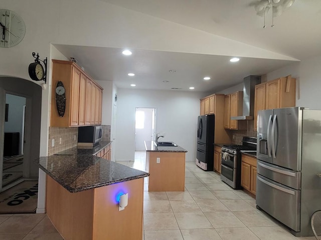 kitchen with light tile patterned flooring, a center island with sink, dark stone countertops, appliances with stainless steel finishes, and wall chimney range hood