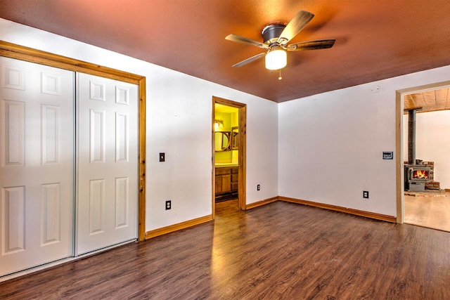 unfurnished bedroom with dark wood-type flooring, a wood stove, a closet, ceiling fan, and ensuite bath