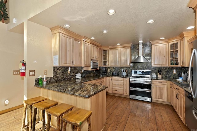 kitchen featuring kitchen peninsula, stainless steel appliances, wall chimney range hood, and wood-type flooring