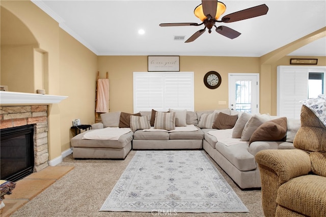 carpeted living room with a stone fireplace, ornamental molding, and ceiling fan