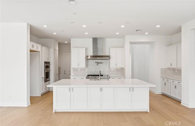 kitchen featuring white cabinetry, wall chimney exhaust hood, backsplash, an island with sink, and light hardwood / wood-style floors