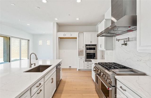 kitchen featuring wall chimney range hood, sink, light stone counters, white cabinetry, and stainless steel appliances