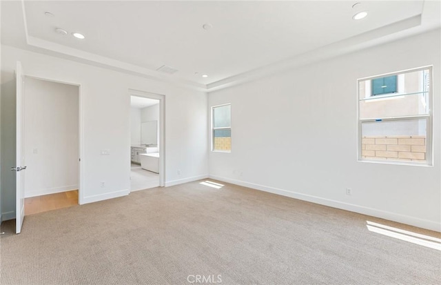 unfurnished bedroom featuring a raised ceiling, light colored carpet, connected bathroom, and multiple windows