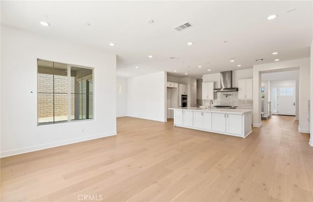 kitchen with sink, wall chimney range hood, light hardwood / wood-style flooring, white cabinets, and an island with sink