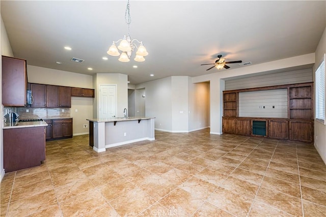 kitchen with ceiling fan with notable chandelier, pendant lighting, a breakfast bar area, backsplash, and a center island with sink