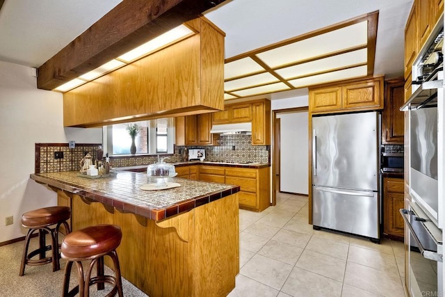 kitchen featuring tile countertops, kitchen peninsula, backsplash, a breakfast bar, and appliances with stainless steel finishes