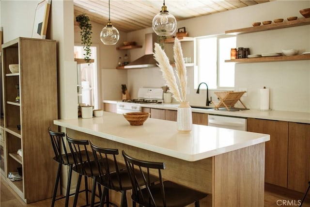 kitchen featuring white appliances, decorative light fixtures, wall chimney range hood, sink, and a breakfast bar