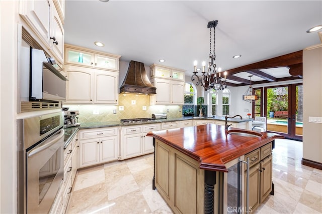 kitchen featuring appliances with stainless steel finishes, an island with sink, pendant lighting, custom range hood, and beam ceiling