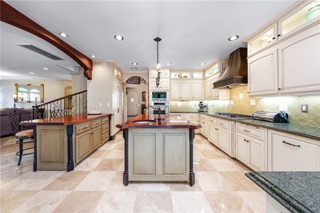 kitchen with appliances with stainless steel finishes, cream cabinetry, pendant lighting, custom range hood, and a center island with sink