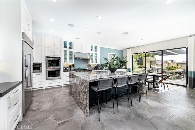 kitchen featuring a kitchen breakfast bar, stainless steel double oven, exhaust hood, white cabinetry, and an island with sink