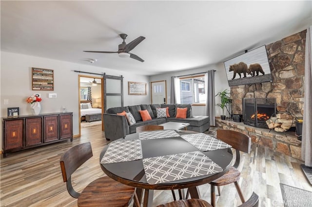 dining room featuring a barn door, ceiling fan, light hardwood / wood-style flooring, and a stone fireplace