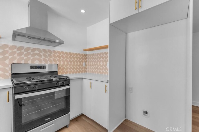 kitchen with wall chimney range hood, light wood-type flooring, backsplash, stainless steel range with gas stovetop, and white cabinetry