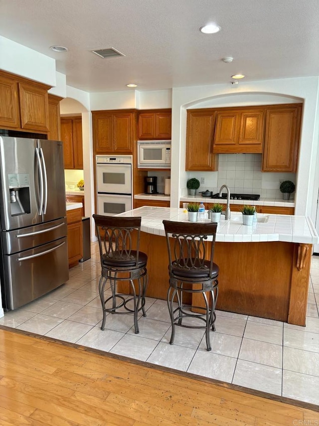 kitchen featuring white appliances, a center island with sink, light hardwood / wood-style flooring, tile counters, and a kitchen bar