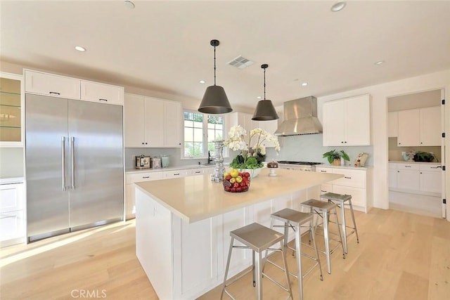 kitchen with built in fridge, light hardwood / wood-style flooring, wall chimney exhaust hood, a kitchen island, and white cabinetry