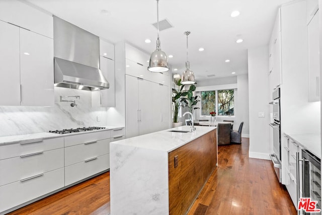 kitchen with hanging light fixtures, wine cooler, white cabinets, a kitchen island with sink, and sink