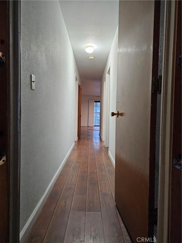 hall with a textured ceiling, a textured wall, wood-type flooring, and baseboards