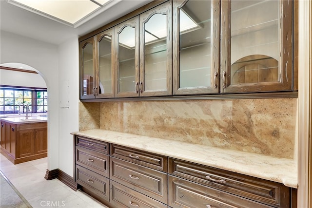 kitchen with light tile patterned floors, decorative backsplash, dark brown cabinets, light stone countertops, and sink