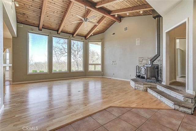 unfurnished living room featuring beamed ceiling, a wood stove, ceiling fan, wooden ceiling, and light wood-type flooring