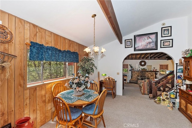 carpeted dining space featuring lofted ceiling with beams, a stone fireplace, wooden walls, and a chandelier