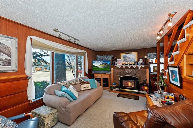 carpeted living room featuring a textured ceiling, a stone fireplace, wooden walls, and track lighting