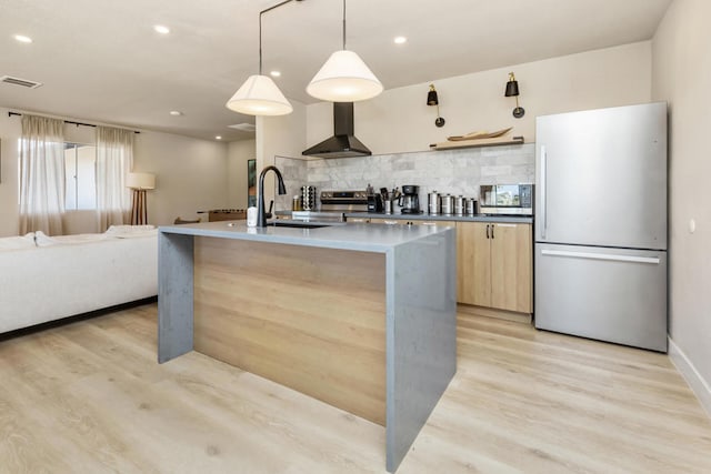 kitchen with an island with sink, decorative light fixtures, wall chimney range hood, stainless steel appliances, and light hardwood / wood-style floors