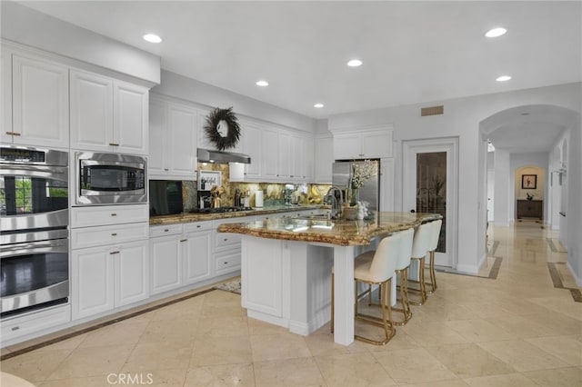 kitchen with range hood, a center island with sink, light stone countertops, appliances with stainless steel finishes, and white cabinets