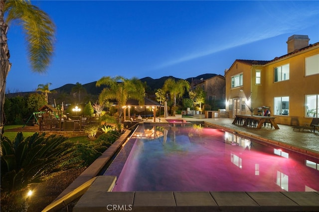 pool at dusk with a gazebo, a patio area, a mountain view, and a playground