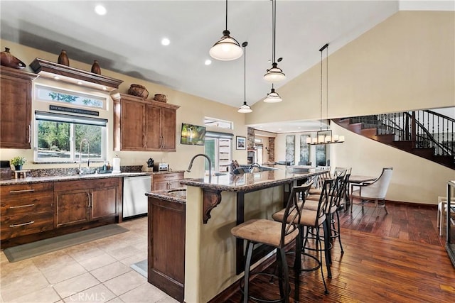 kitchen featuring a kitchen bar, a center island with sink, decorative light fixtures, dark stone counters, and stainless steel dishwasher