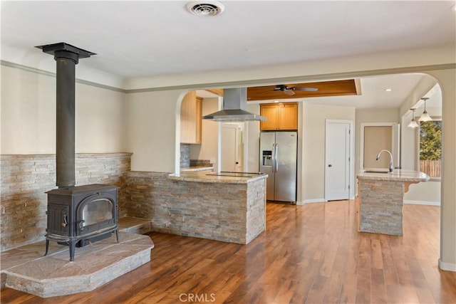 kitchen featuring wood finished floors, visible vents, a wood stove, wall chimney range hood, and stainless steel fridge