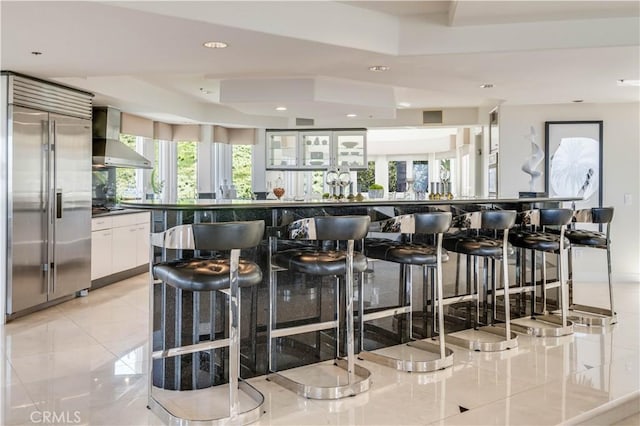 kitchen with light tile patterned flooring, wall chimney exhaust hood, appliances with stainless steel finishes, a kitchen breakfast bar, and white cabinets