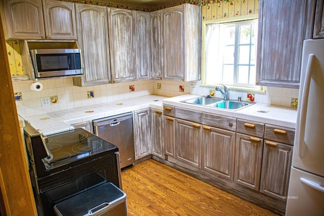 kitchen with tile counters, sink, stainless steel appliances, backsplash, and light hardwood / wood-style floors