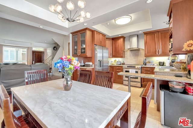 kitchen featuring appliances with stainless steel finishes, an inviting chandelier, a raised ceiling, decorative light fixtures, and wall chimney exhaust hood