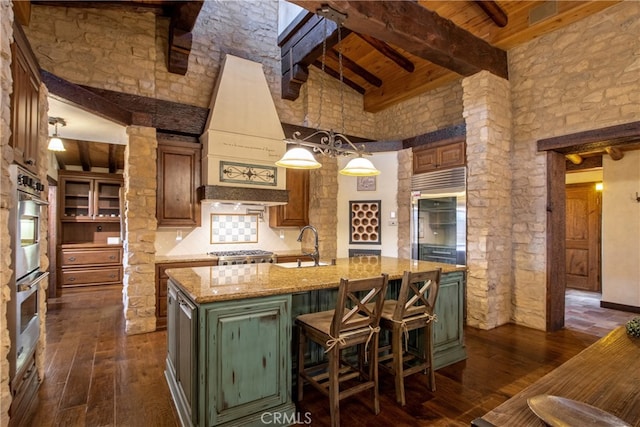 kitchen with dark wood-type flooring, beam ceiling, high vaulted ceiling, a kitchen island with sink, and light stone countertops