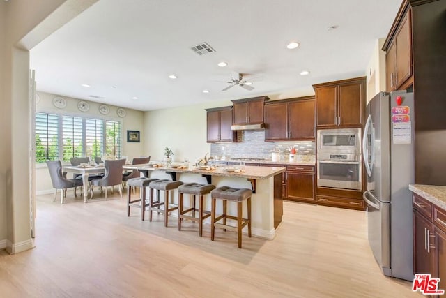 kitchen with appliances with stainless steel finishes, light wood-type flooring, a center island with sink, a kitchen breakfast bar, and light stone countertops