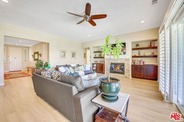 living room with built in features, a stone fireplace, light hardwood / wood-style flooring, and ceiling fan