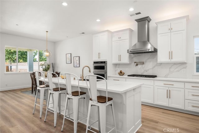 kitchen with light wood-type flooring, white cabinetry, a kitchen island with sink, and wall chimney range hood