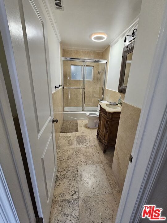 full bathroom featuring bath / shower combo with glass door, vanity, crown molding, tile walls, and toilet