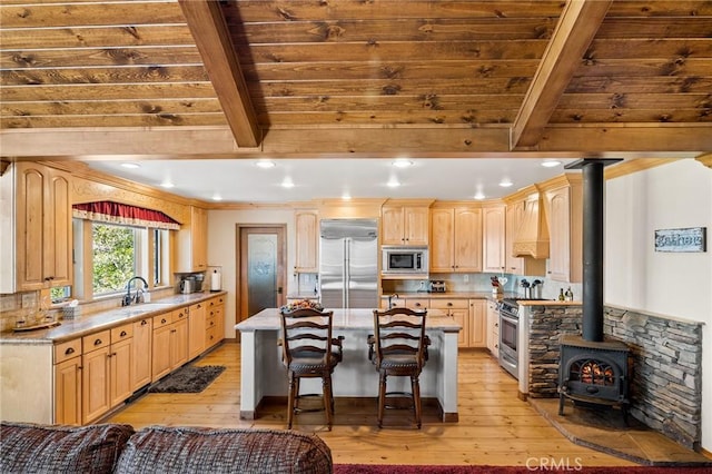 kitchen with a wood stove, built in appliances, beam ceiling, light brown cabinetry, and light hardwood / wood-style floors