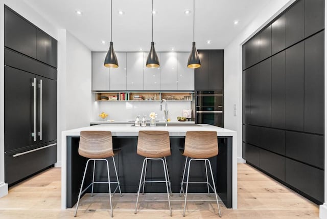 kitchen featuring sink, double oven, paneled built in fridge, decorative light fixtures, and a large island with sink