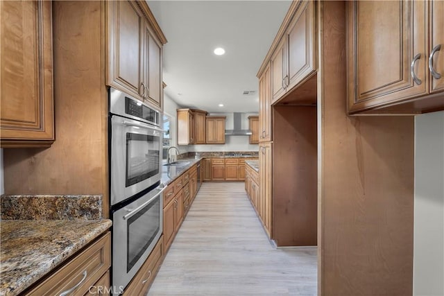 kitchen with wall chimney exhaust hood, double oven, dark stone countertops, sink, and light hardwood / wood-style flooring