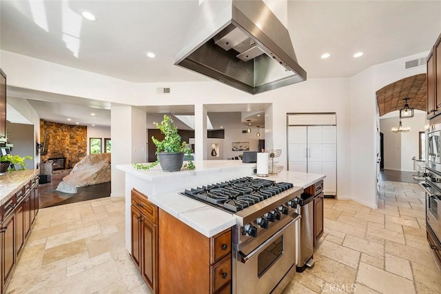 kitchen with appliances with stainless steel finishes, an inviting chandelier, a center island, a fireplace, and wall chimney exhaust hood