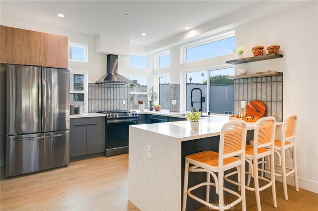 kitchen with light hardwood / wood-style flooring, appliances with stainless steel finishes, wall chimney range hood, sink, and a breakfast bar area