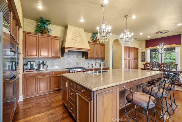 kitchen with decorative light fixtures, dark wood-type flooring, premium range hood, light stone counters, and a center island with sink