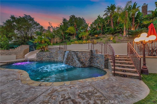 pool at dusk with pool water feature, a water slide, and a patio