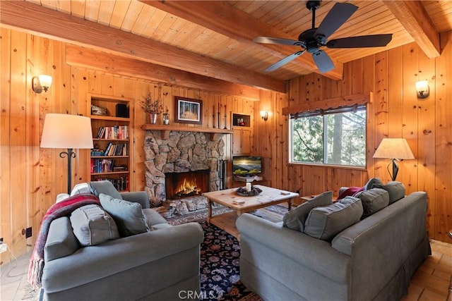 living room with ceiling fan, beam ceiling, a stone fireplace, and wooden walls