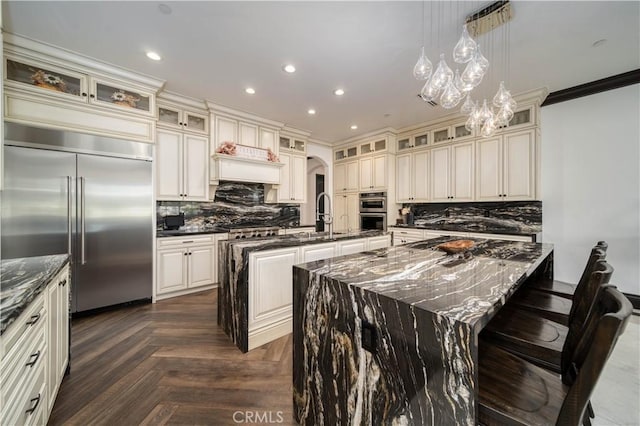 kitchen with sink, appliances with stainless steel finishes, dark parquet floors, decorative light fixtures, and a large island with sink