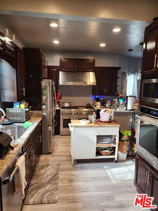 kitchen with dark brown cabinetry, appliances with stainless steel finishes, wall chimney exhaust hood, decorative light fixtures, and light hardwood / wood-style flooring