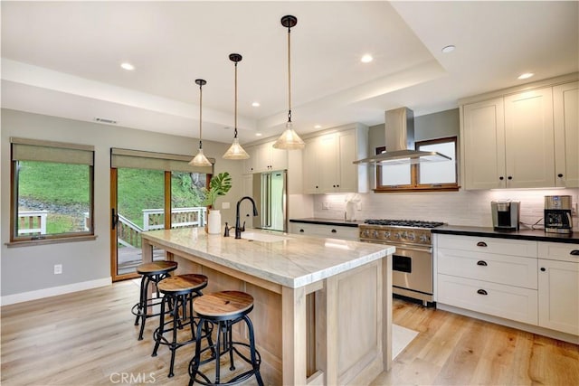 kitchen featuring an island with sink, dark stone countertops, range hood, a raised ceiling, and high quality appliances